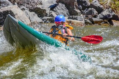 Whitewater kayaker in a green inflatable kayak navigating rocky river rapids, wearing a blue helmet, orange life jacket and pink sunglasses, paddling with a red-bladed paddle through splashing whitewater.