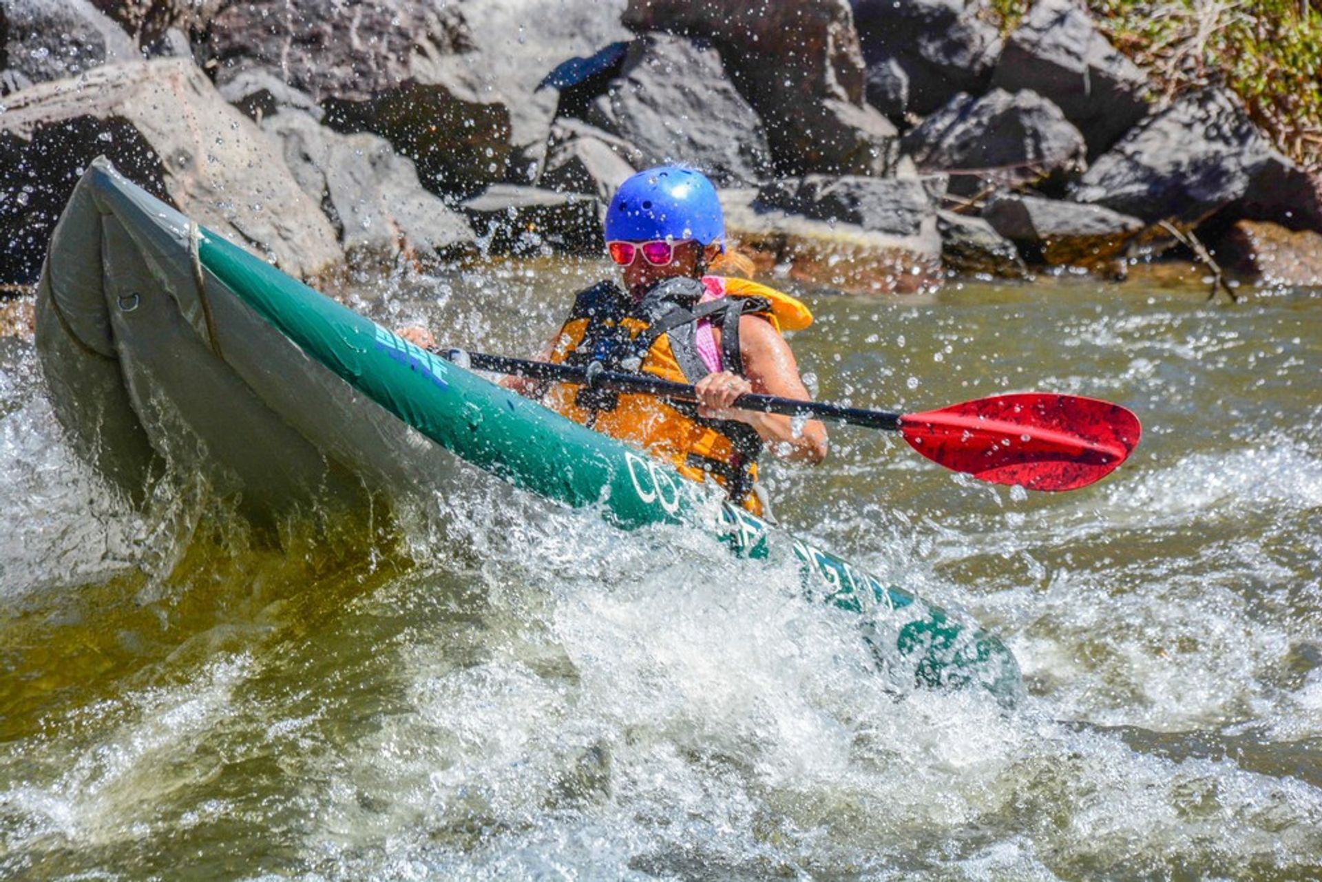 Whitewater kayaker in a green inflatable kayak navigating rocky river rapids, wearing a blue helmet, orange life jacket and pink sunglasses, paddling with a red-bladed paddle through splashing whitewater.
