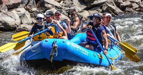 Group of six paddlers in a bright blue inflatable raft navigating whitewater on a rocky mountain river, wearing life jackets and hats and energetically paddling yellow oars — outdoor whitewater rafting adventure.