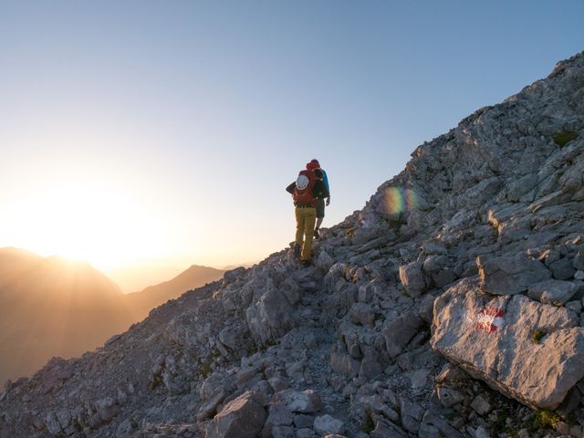Two hikers with backpacks climbing a rocky mountain ridge at sunrise, golden light over distant peaks and a red-white trail marker on limestone