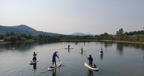 Group of stand-up paddleboarders on a calm mountain lake with tree-lined shore, rolling hills and distant peaks