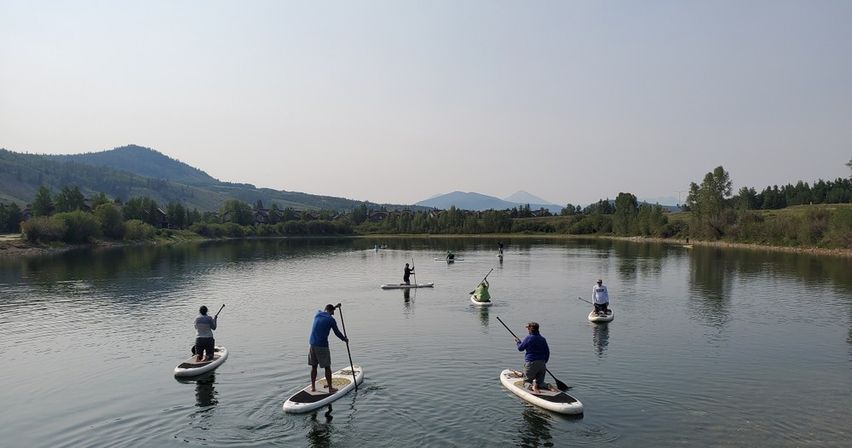 Group of stand-up paddleboarders on a calm mountain lake with tree-lined shore, rolling hills and distant peaks