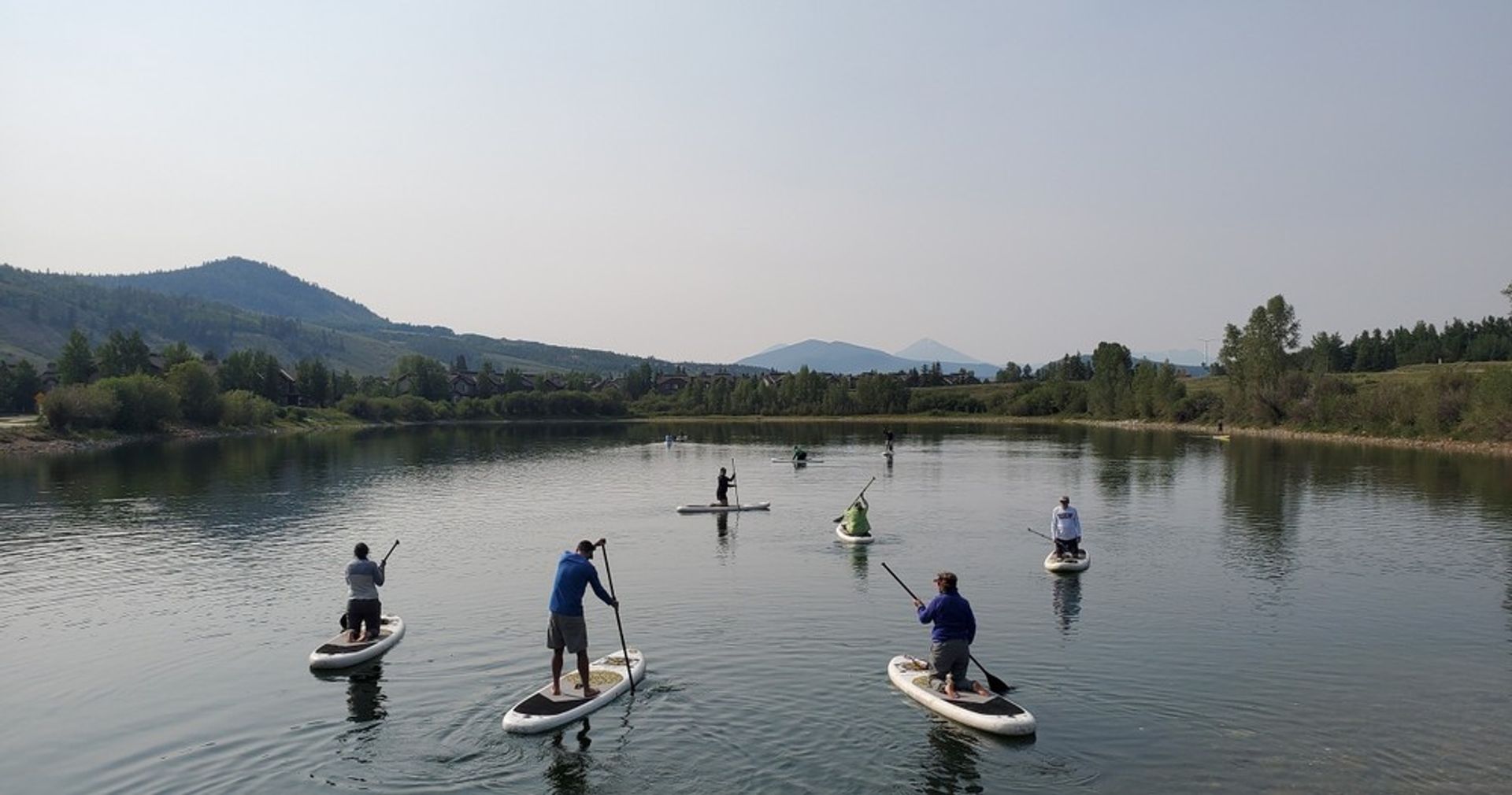 Group of stand-up paddleboarders on a calm mountain lake with tree-lined shore, rolling hills and distant peaks
