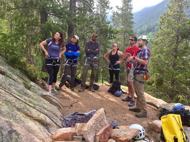 Six climbers in harnesses gather on a rocky alpine ledge in a pine forest as an instructor gestures, with ropes, helmets, and backpacks scattered on the ground.
