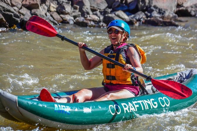 Smiling kayaker in a blue helmet and yellow life jacket paddles a green inflatable kayak with red blades through rocky river rapids.