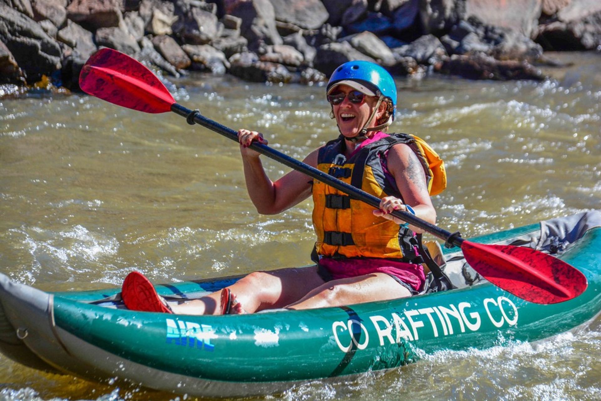 Smiling kayaker in a blue helmet and yellow life jacket paddles a green inflatable kayak with red blades through rocky river rapids.