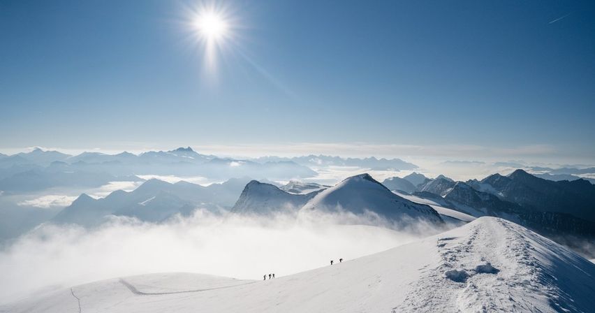 Sunlit alpine ridge under a clear blue sky, snow-covered peaks and low clouds filling the valleys, with a tiny group of hikers trekking along the snowy summit path.