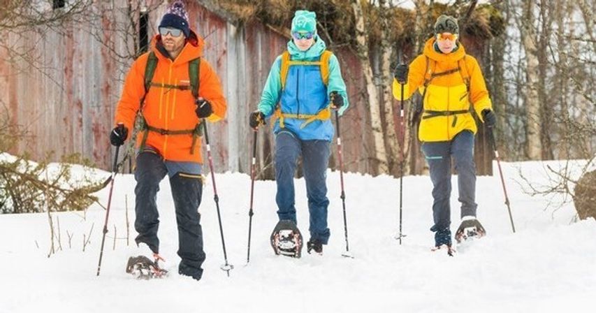 Three people snowshoeing on a snow-covered trail through a birch forest past a weathered barn, wearing bright orange, blue, and yellow winter jackets and using trekking poles