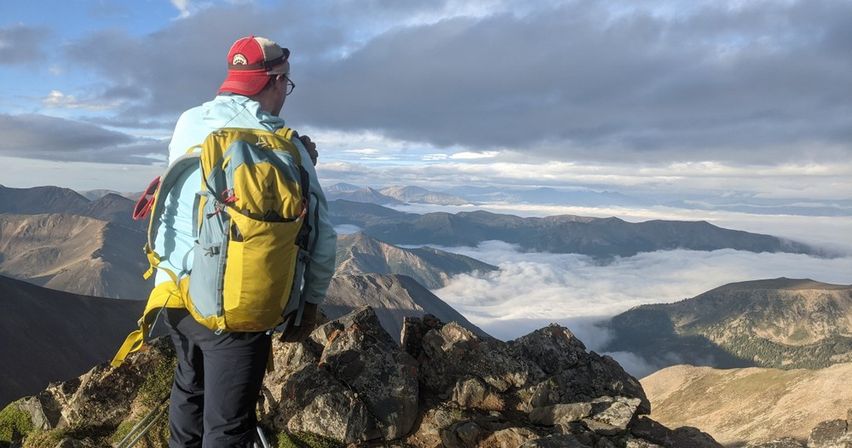 Hiker with yellow backpack and red cap atop a rocky alpine summit, surveying a sea of clouds filling the mountain valley and layered distant ridgelines.