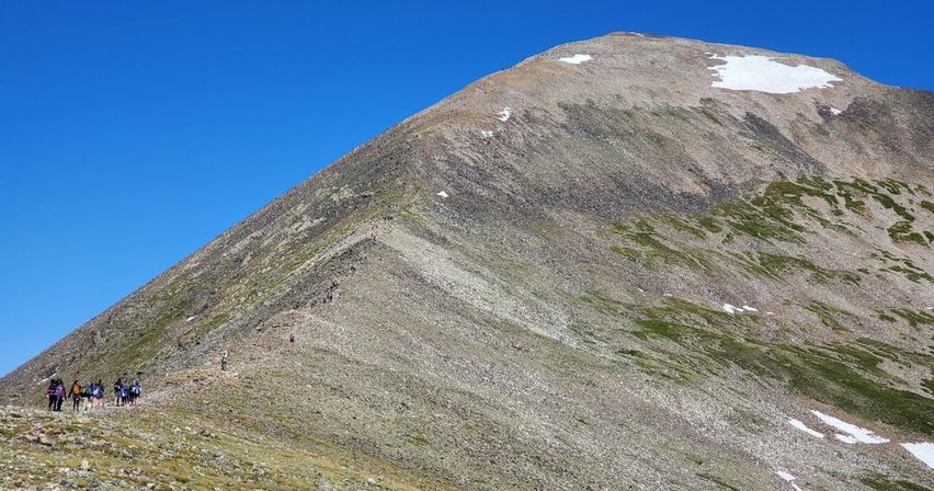 Small group of hikers trekking along a rocky alpine ridge toward a snow-dusted summit under a bright clear blue sky.