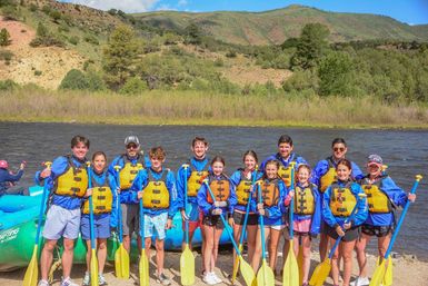Group of rafters in yellow life jackets and blue jackets holding yellow paddles beside a blue inflatable raft on a sunny mountain river with green hills