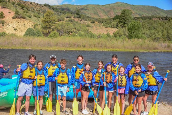 Group of rafters in yellow life jackets and blue jackets holding yellow paddles beside a blue inflatable raft on a sunny mountain river with green hills