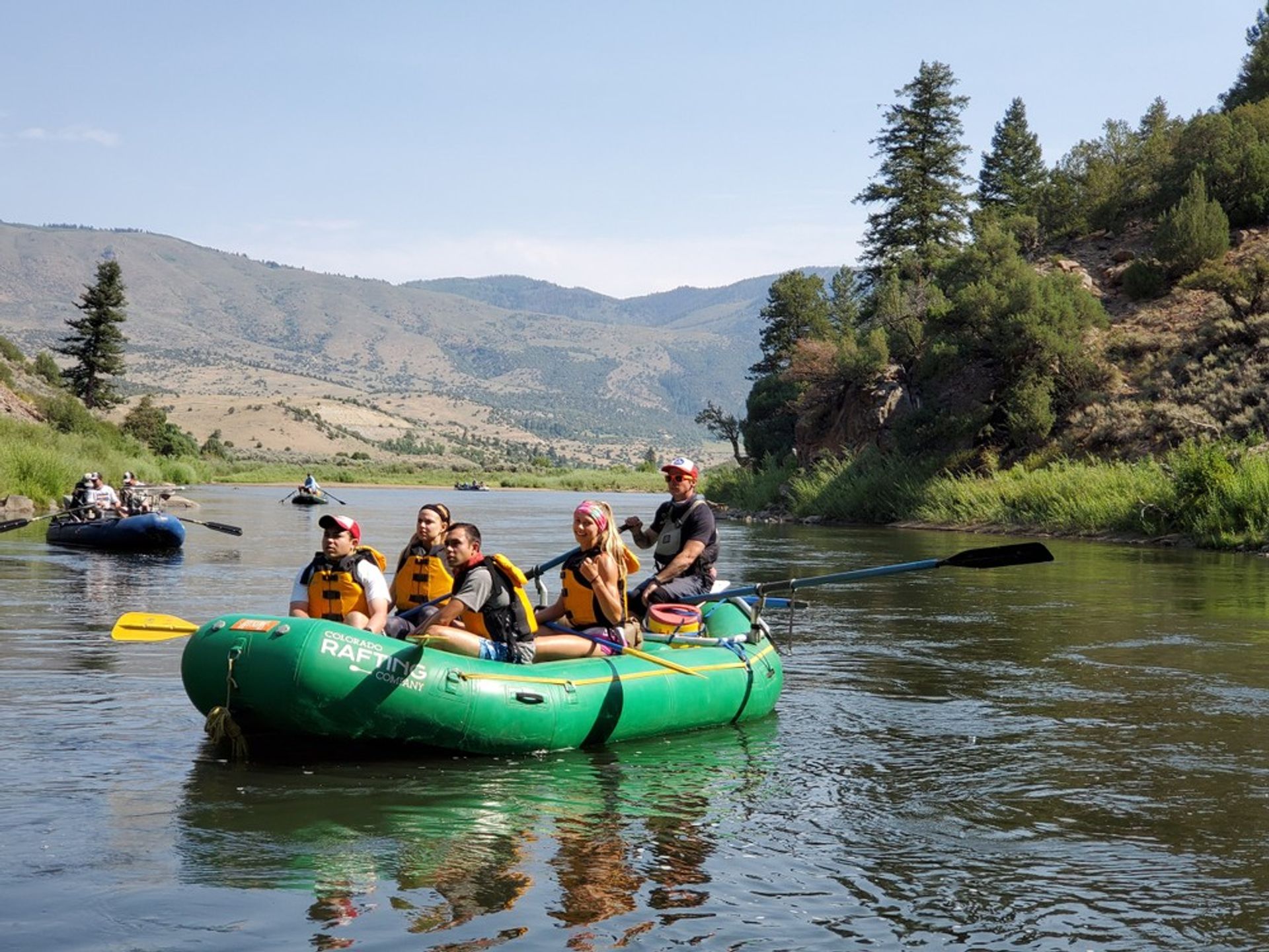 Group in yellow life jackets on a green inflatable raft enjoying river rafting on a sunny mountain river with pine-covered banks.