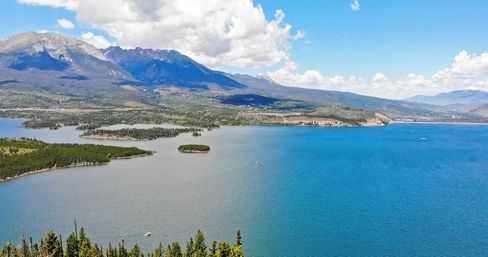 Aerial panoramic view of a sparkling deep-blue alpine lake with small pine-covered islands, forested shorelines and rugged mountain peaks under a bright blue sky with fluffy clouds.