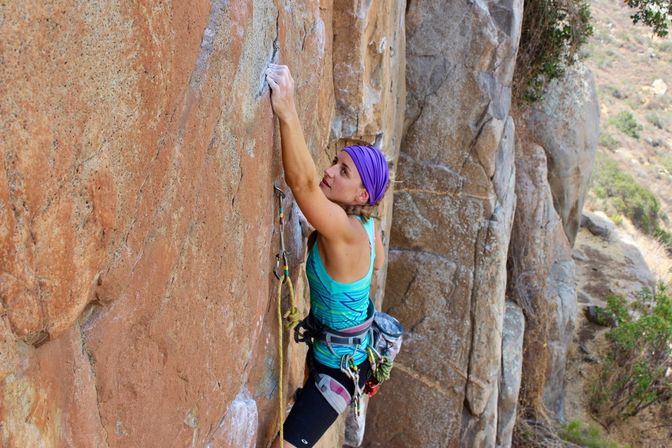 Female rock climber in a purple headband and teal tank top scaling a sunlit orange-brown cliff face, clipped into rope and quickdraws with a scrubby hillside below.