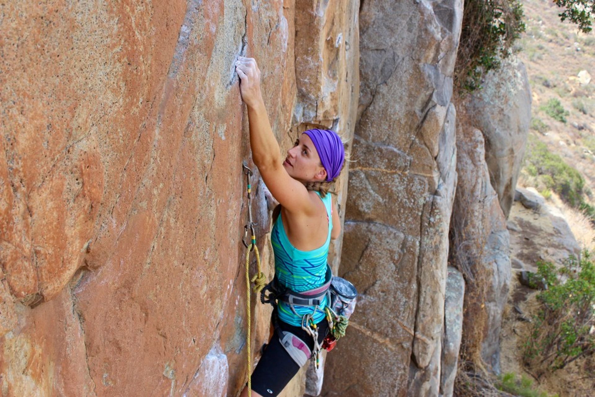 Female rock climber in a purple headband and teal tank top scaling a sunlit orange-brown cliff face, clipped into rope and quickdraws with a scrubby hillside below.
