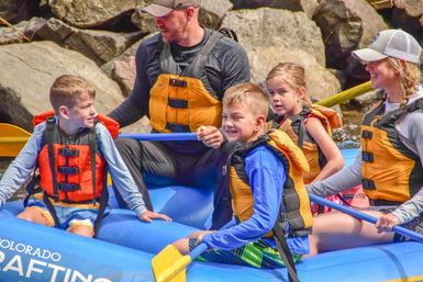Family group wearing life jackets smiling on a blue inflatable raft while river rafting along rocky banks in Colorado
