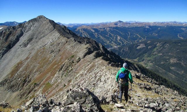 Backpacker in a green jacket using trekking poles traverses a rocky alpine ridge with panoramic mountain peaks and forested valleys under a clear blue sky.