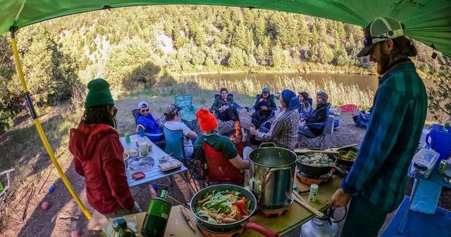 Lakeside forest campsite with a group gathered around a campfire, portable stoves and pans cooking colorful vegetables under a green tarp