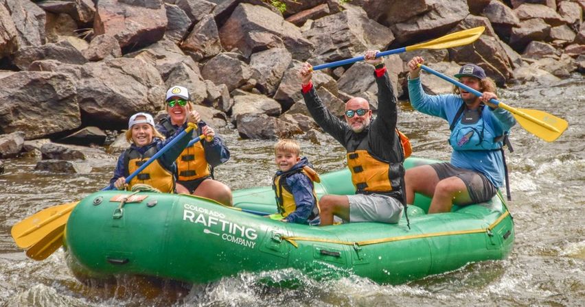 Five rafters in bright life jackets cheer and raise paddles in a green inflatable on a rocky Colorado whitewater river