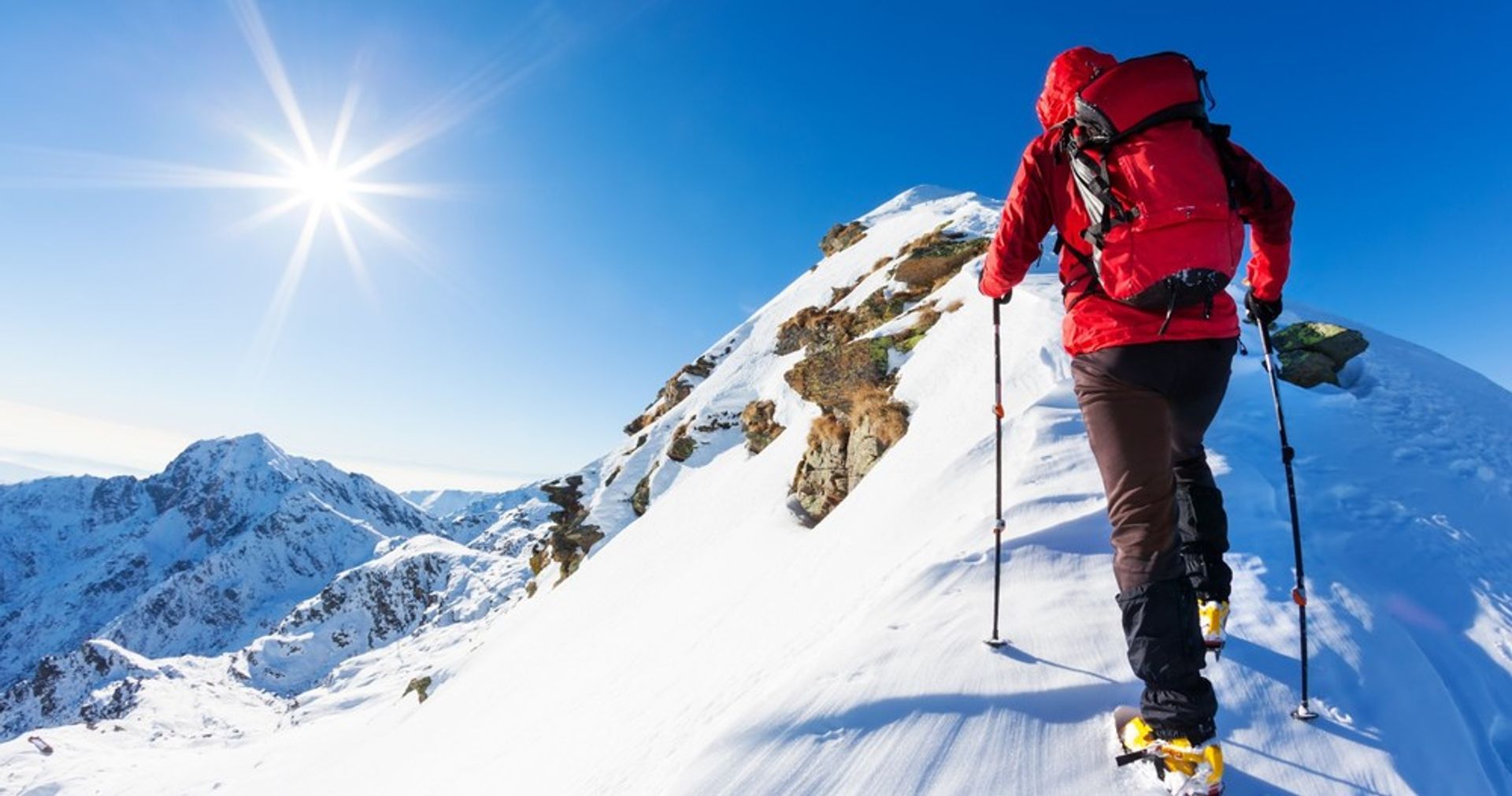 Backcountry hiker in a red jacket with backpack and trekking poles ascending a sunny, snow-covered alpine ridge with rugged peaks under a clear blue sky