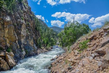 Whitewater rafting on a fast mountain river cutting through a rocky canyon, surrounded by pine trees and a bright blue sky.