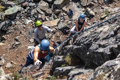 Three outdoor rock climbers on a rugged mountainside: a climber in a blue helmet reaches up a steep rock face while a belayer with a neon green helmet manages ropes and a partner waits on the boulder-strewn ledge.