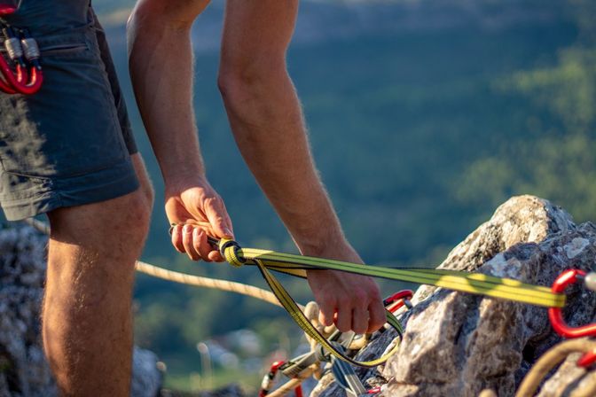 Close-up of a rock climber securing a yellow sling and multiple carabiners to a rock anchor, legs visible and sunlit mountain valley in the background