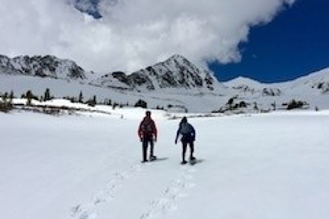 Two people snowshoeing across a wide snowy alpine valley toward jagged mountain peaks under a blue sky with clouds