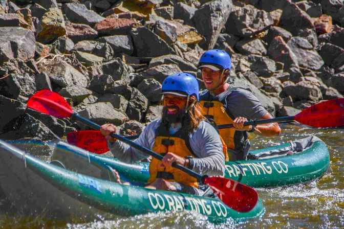 River kayaking: two people in blue helmets and yellow life jackets paddle green inflatable kayaks with red paddles along a rocky riverbank.