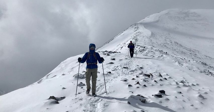 Two hikers with trekking poles on a snow-covered alpine ridge, footprints trailing behind them across rocky snow fields beneath a cloudy gray sky