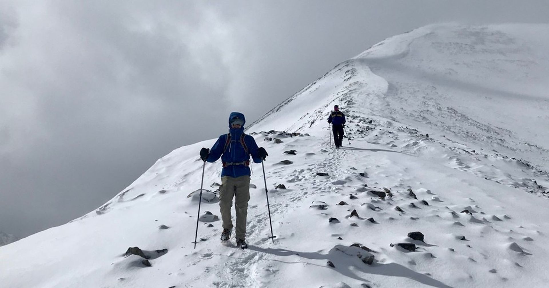 Two hikers with trekking poles on a snow-covered alpine ridge, footprints trailing behind them across rocky snow fields beneath a cloudy gray sky