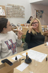 Two people in a craft studio sniffing scent strips during a perfume-making workshop, table with bottles, notes and a workshop sign in the background.
