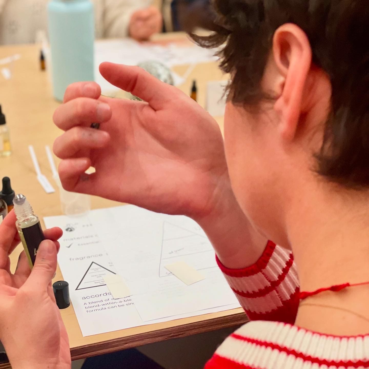 Participant at a perfume workshop smelling a roller bottle on their wrist over a table with fragrance strips, droppers and a scent pyramid worksheet.
