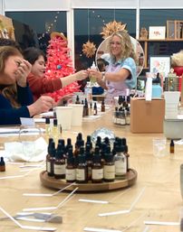 Participants in a cozy artisan studio making fragrances — trays of amber dropper bottles and scent strips on a table, instructor handing a bottle near a pink holiday tree.