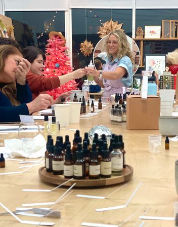 Participants in a cozy artisan studio making fragrances — trays of amber dropper bottles and scent strips on a table, instructor handing a bottle near a pink holiday tree.