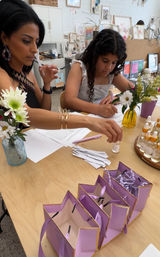 Adult and child at a bright art studio table doing a DIY scent workshop — sniffing fragrance strips, filling forms, tiny glass vials, fresh flowers in vases and a row of purple gift bags on a wooden table.
