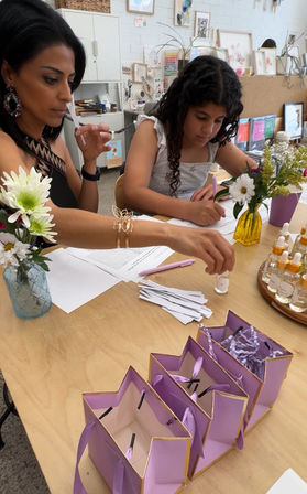 Adult and child at a bright art studio table doing a DIY scent workshop — sniffing fragrance strips, filling forms, tiny glass vials, fresh flowers in vases and a row of purple gift bags on a wooden table.