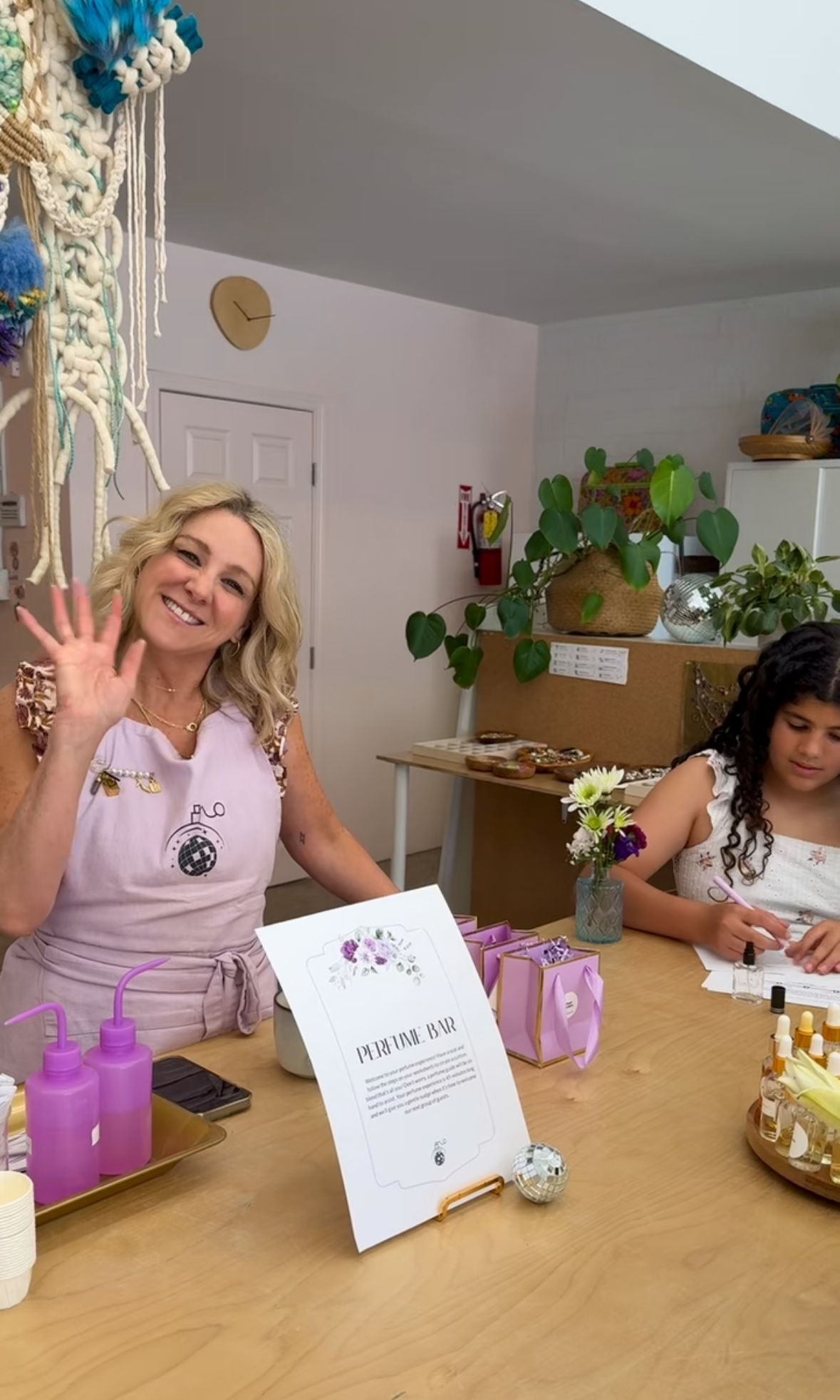 Smiling woman waving at a DIY perfume bar workshop — purple squeeze bottles, gift boxes and a girl mixing scents at a wooden table in a bright, plant-filled studio with macramé wall art.
