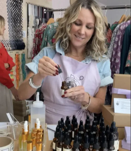Smiling woman in an apron fills an amber dropper bottle with a pipette, surrounded by rows of small-batch skincare serums and gold-capped bottles on a boutique counter with a colorful clothing rack behind her.