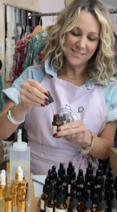 Smiling woman in an apron fills an amber dropper bottle with a pipette, surrounded by rows of small-batch skincare serums and gold-capped bottles on a boutique counter with a colorful clothing rack behind her.