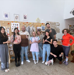Smiling group in a bright indoor DIY craft workshop holding handmade ornaments and tools, posed in front of a display wall reading "Crafting is self care," with a small dog at their feet.