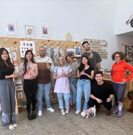Smiling group in a bright indoor DIY craft workshop holding handmade ornaments and tools, posed in front of a display wall reading "Crafting is self care," with a small dog at their feet.