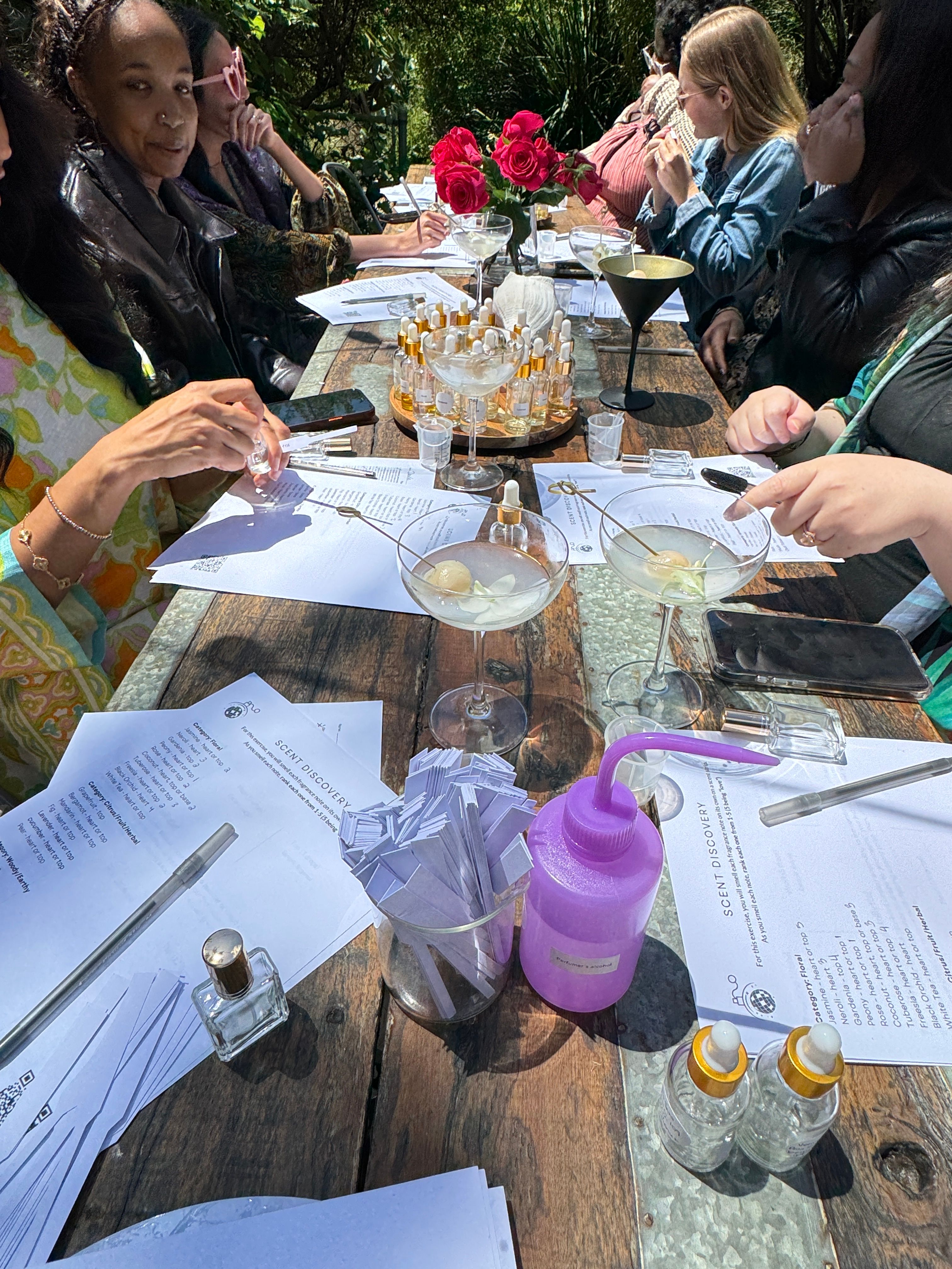 Group of people at an outdoor scent-discovery workshop around a rustic wooden table with perfume sample vials, paper scent strips in a jar, printed worksheets, cocktail glasses with garnishes, a purple squeeze bottle and red roses centerpiece.
