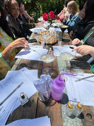 Group of people at an outdoor scent-discovery workshop around a rustic wooden table with perfume sample vials, paper scent strips in a jar, printed worksheets, cocktail glasses with garnishes, a purple squeeze bottle and red roses centerpiece.
