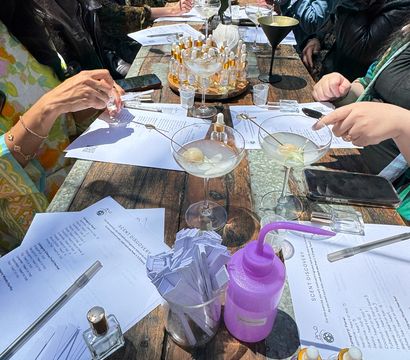 Group of people at an outdoor scent-discovery workshop around a rustic wooden table with perfume sample vials, paper scent strips in a jar, printed worksheets, cocktail glasses with garnishes, a purple squeeze bottle and red roses centerpiece.