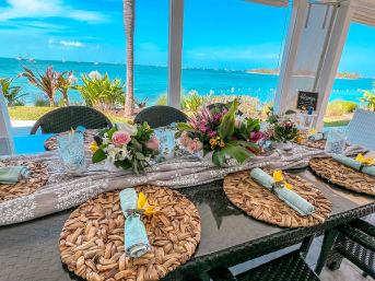 Sunny tropical ocean-view patio dining table with woven placemats, pastel napkins topped with yellow orchids and lush floral centerpieces, palm tree and sailboats on turquoise water