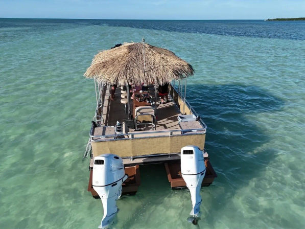 Tiki-style pontoon boat with a thatched roof and twin outboard engines floating on clear turquoise tropical shallow sea under a bright blue sky
