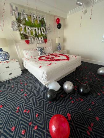 Romantic hotel room birthday setup: white bed with red rose-petal heart, silver "HAPPY BIRTHDAY" balloons on the headboard and red, black and silver balloons scattered on patterned carpet.
