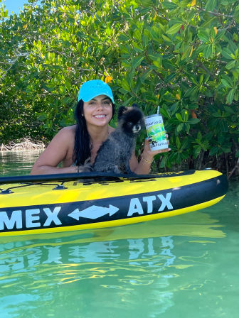 Smiling woman in a turquoise cap on a yellow inflatable kayak in clear tropical mangrove waters, holding a colorful travel cup while a small black fluffy dog perches on the kayak.